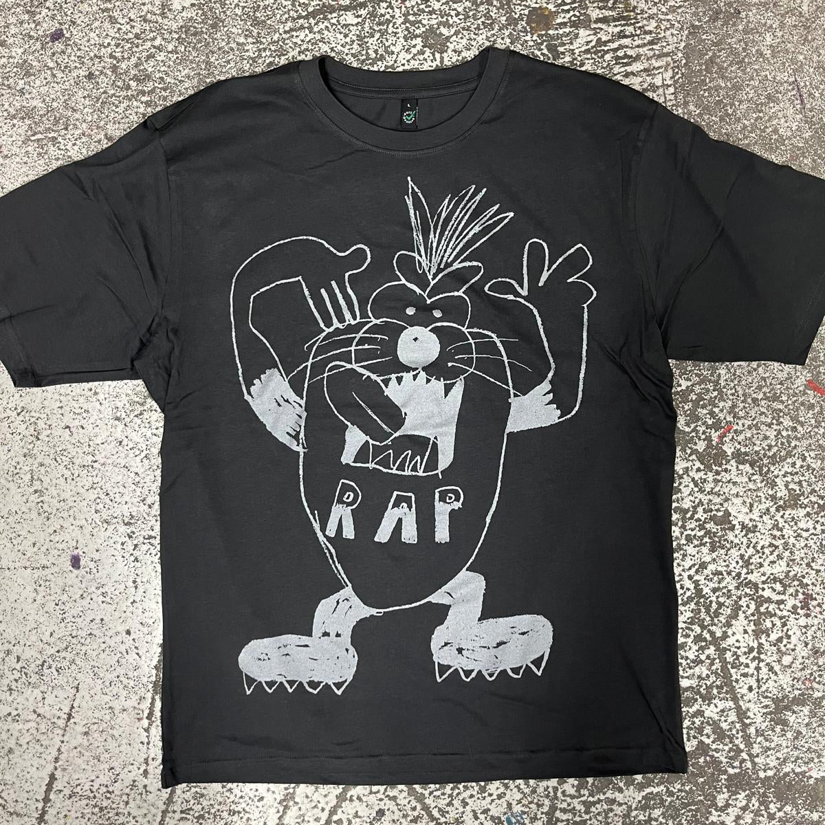 A black short-sleeve graphic t-shirt featuring a white line art print of a cartoon character with the word 'RAP' on its belly, laid flat on a textured concrete floor.